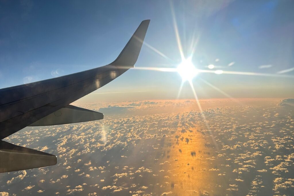 View from a plane window showing the wing, fluffy clouds below, and the sun shining brightly in a clear sky.