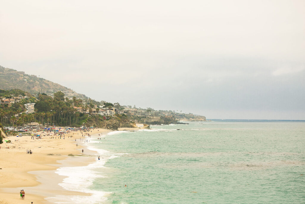 A sandy beach stretches along the coastline with gentle waves. People are scattered on the beach under a cloudy sky. Hills with houses are visible in the background.