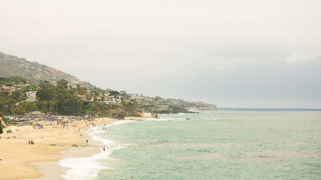 A sandy beach stretches along the coastline with gentle waves. People are scattered on the beach under a cloudy sky. Hills with houses are visible in the background.