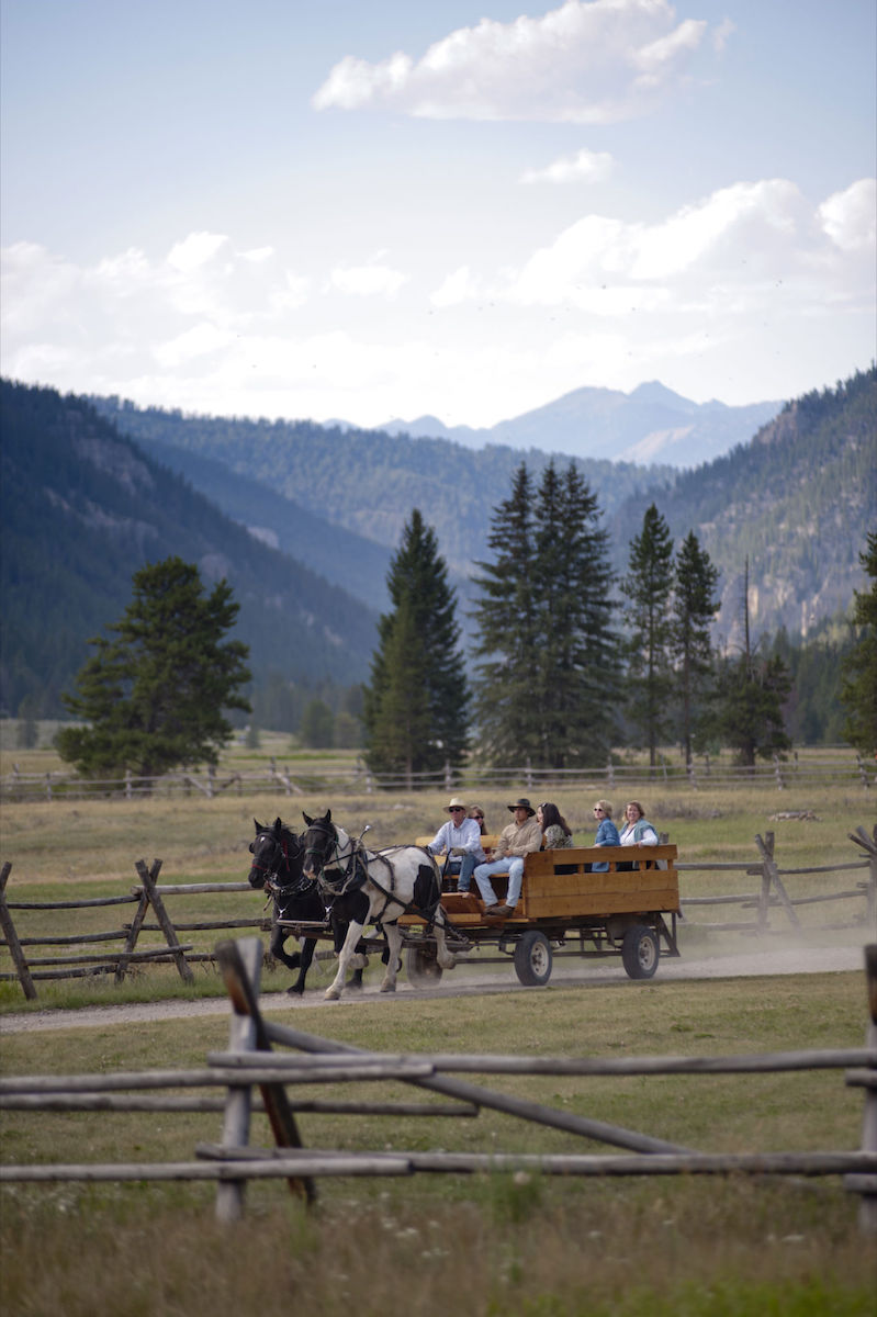 A horse-drawn wagon carrying several people travels along a dirt path in a fenced field with mountains and trees in the background.