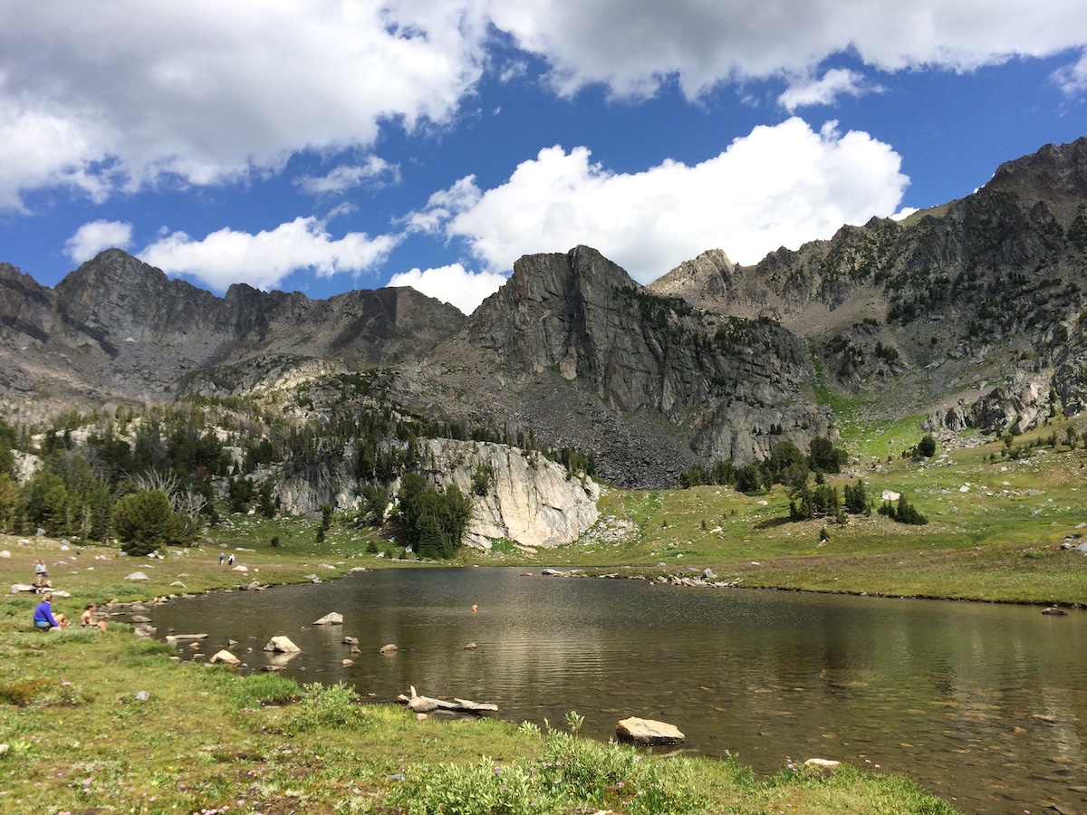 Mountain lake surrounded by grass, rocks, and trees with steep rocky peaks in the background under a partly cloudy sky.