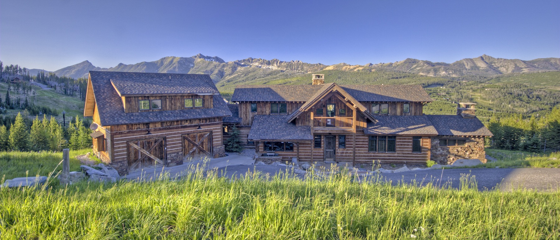 A large wooden lodge-style house with multiple sections sits in a grassy area, surrounded by green hills and mountains under a clear blue sky.