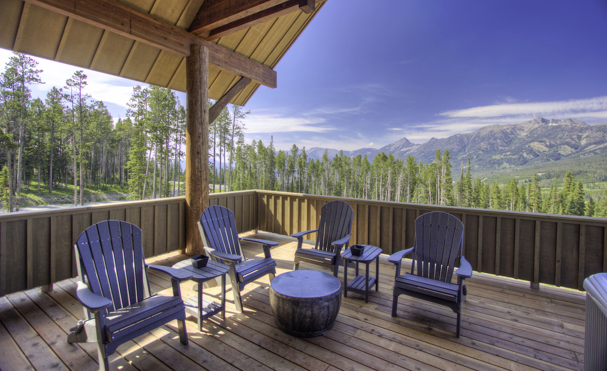 Four Adirondack chairs and a small table sit on a wooden deck under a roof, overlooking a forest and distant mountains under a clear blue sky.