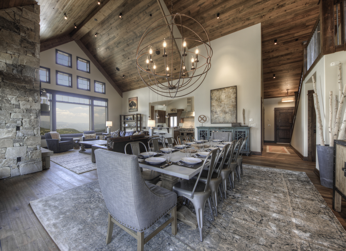 Spacious open-concept dining and living area with vaulted wood ceiling, stone fireplace, large windows, and a long dining table set for eight. Natural light fills the room.