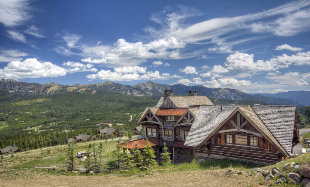 A rustic wooden lodge sits on a hillside with mountains, forests, and scattered houses visible under a partly cloudy blue sky.