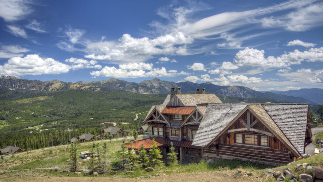 A rustic wooden lodge sits on a hillside with mountains, forests, and scattered houses visible under a partly cloudy blue sky.
