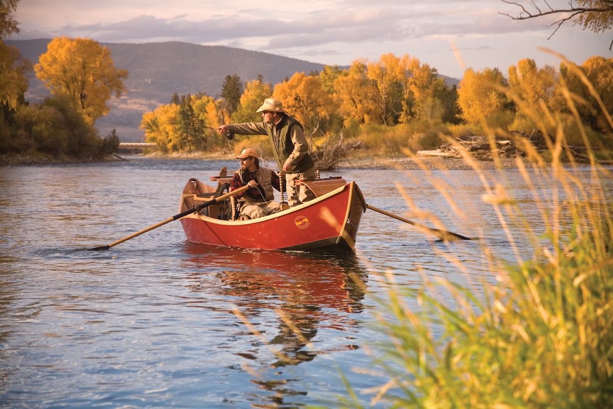 Horizontal fall shot of two men in a red driftboat on the Yellowstone River in Montana. One angler is cating a fly and the other is rowing.