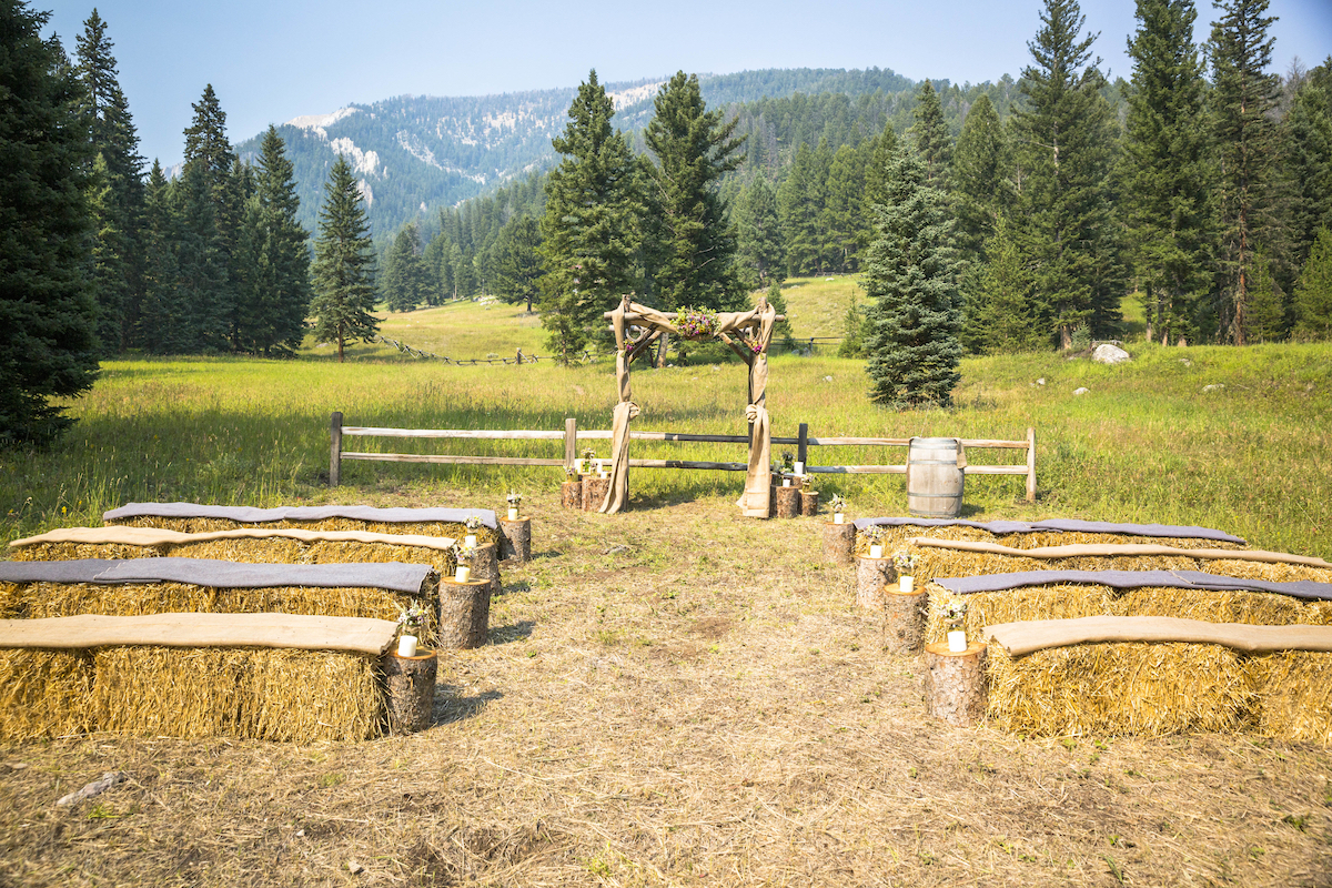Outdoor wedding ceremony setup with hay bale seating, a wooden arch, and forested mountains in the background on a sunny day.