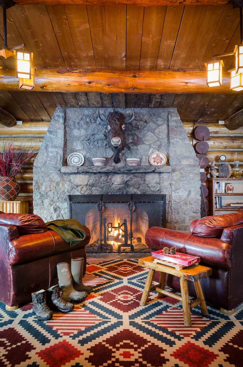 Cozy log cabin living room with stone fireplace, two leather chairs, patterned rug, wooden stool with books, boots on floor, and rustic decor.