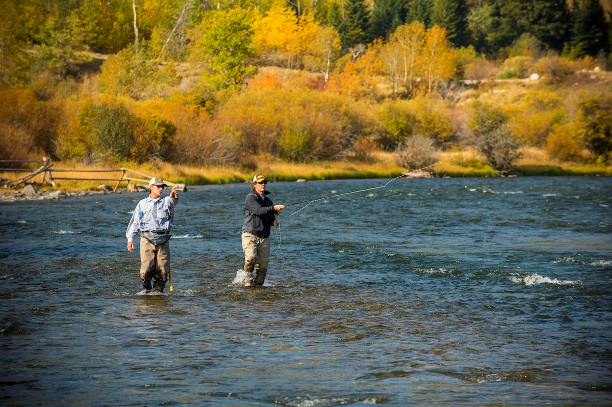 Two people are fly fishing in a river surrounded by autumn trees and colorful foliage.