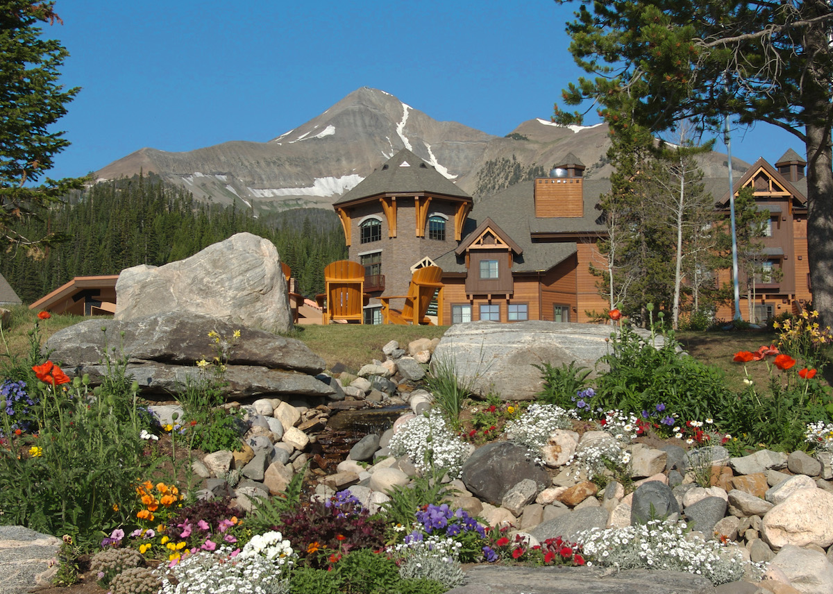 Lodge-style buildings with stone landscaping and colorful flowers in the foreground, with mountains and trees in the background under a clear blue sky.