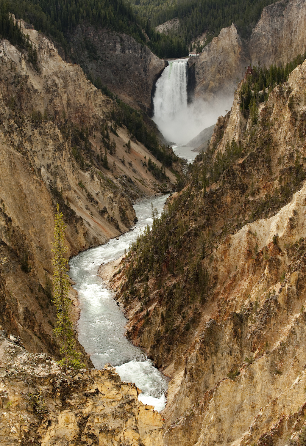 A river flows through a deep canyon with steep rocky walls, leading to a tall waterfall surrounded by forested hills.