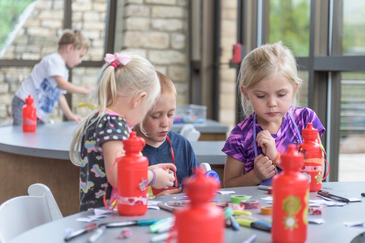 Three young children sit at a table with red water bottles, coloring and crafting; another child stands at a table in the background.
