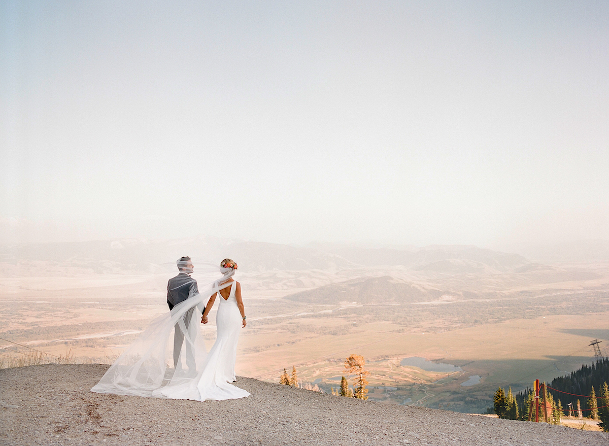 A bride and groom stand together on a mountaintop, holding hands and facing each other, with a long veil and scenic valley in the background.