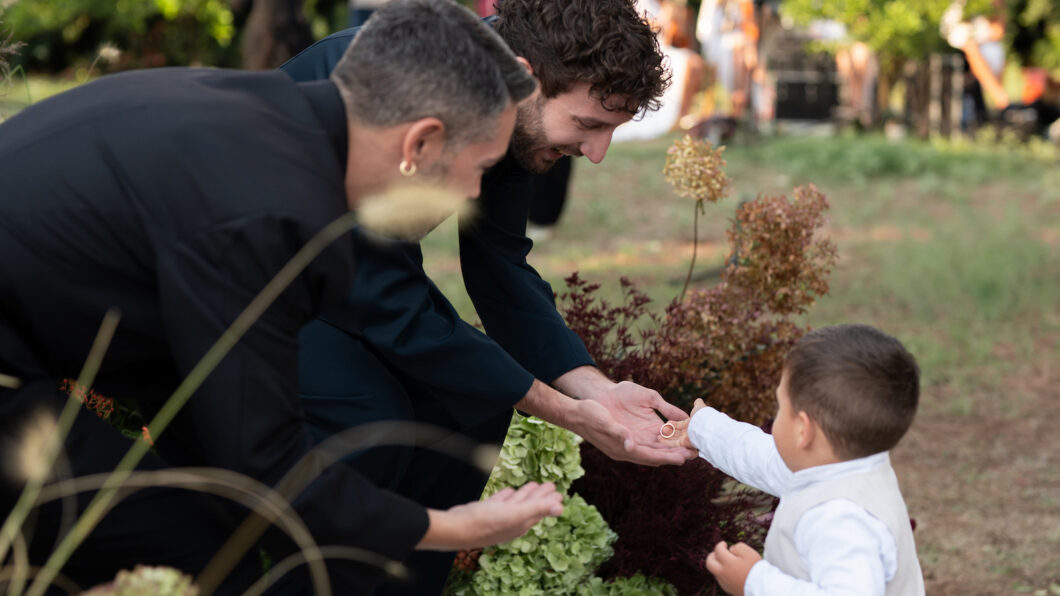 Two adults in dark clothing crouch and reach their hands toward a small child in white, who is standing outdoors among plants.