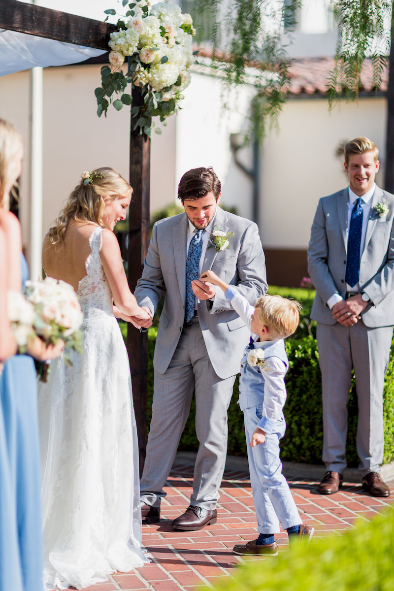 A bride and groom hold hands during an outdoor wedding ceremony, with a young boy in a suit standing beside them and smiling guests in the background.