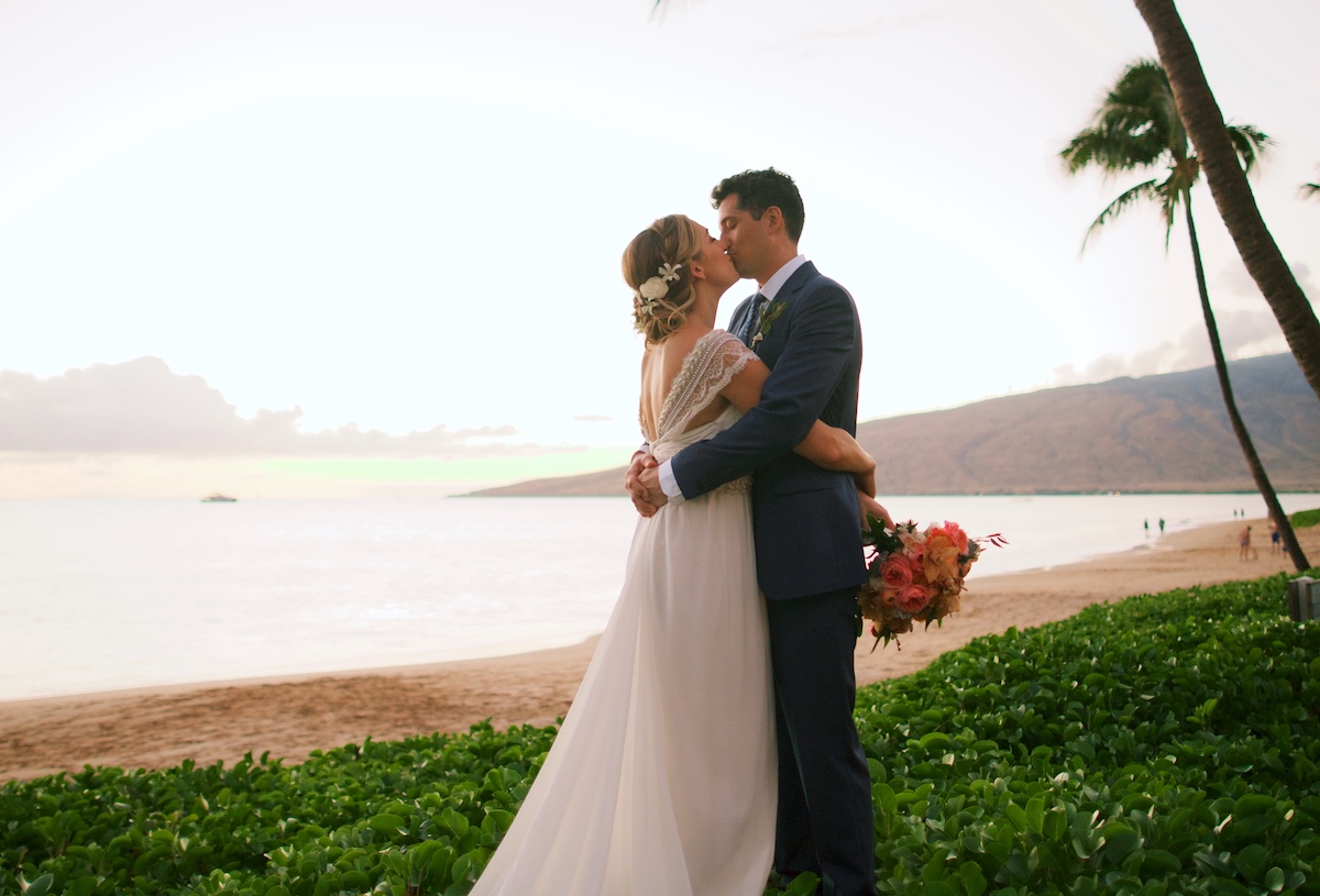 couple embrace on a beach in Maui with greenery and palm trees surrounding