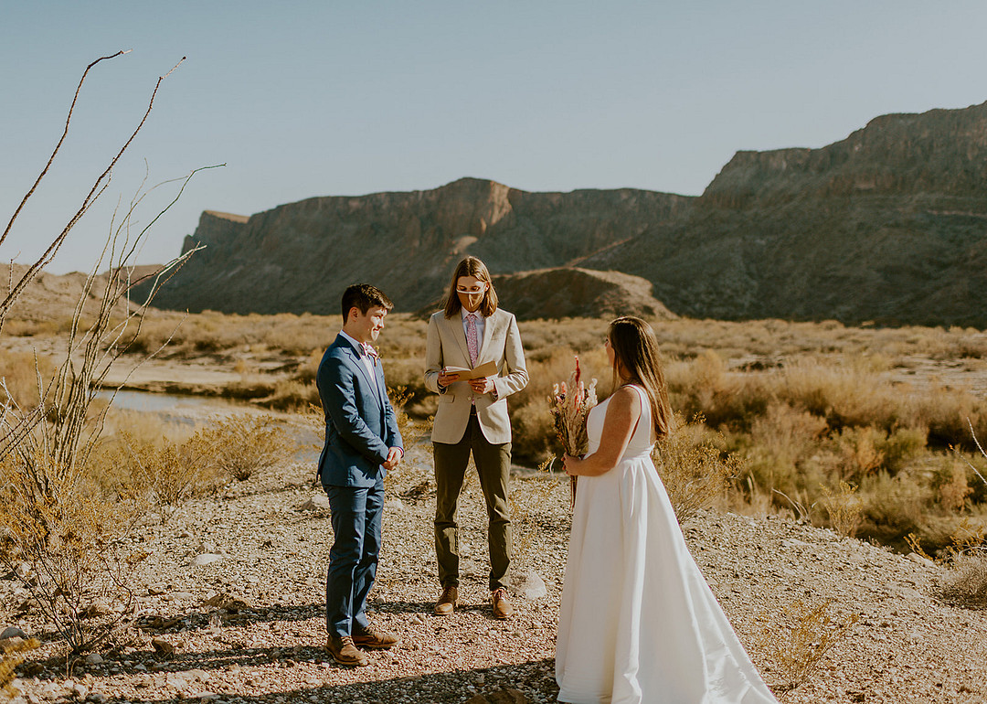 Three people stand in the desert setting of Big Bend National Park; one officiates a wedding ceremony while the other two face each other, dressed in formal attire.