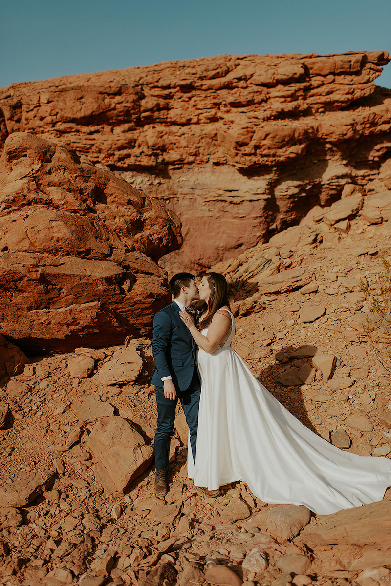 A bride and groom share a kiss while standing on rocky terrain with large red rock formations in the background.