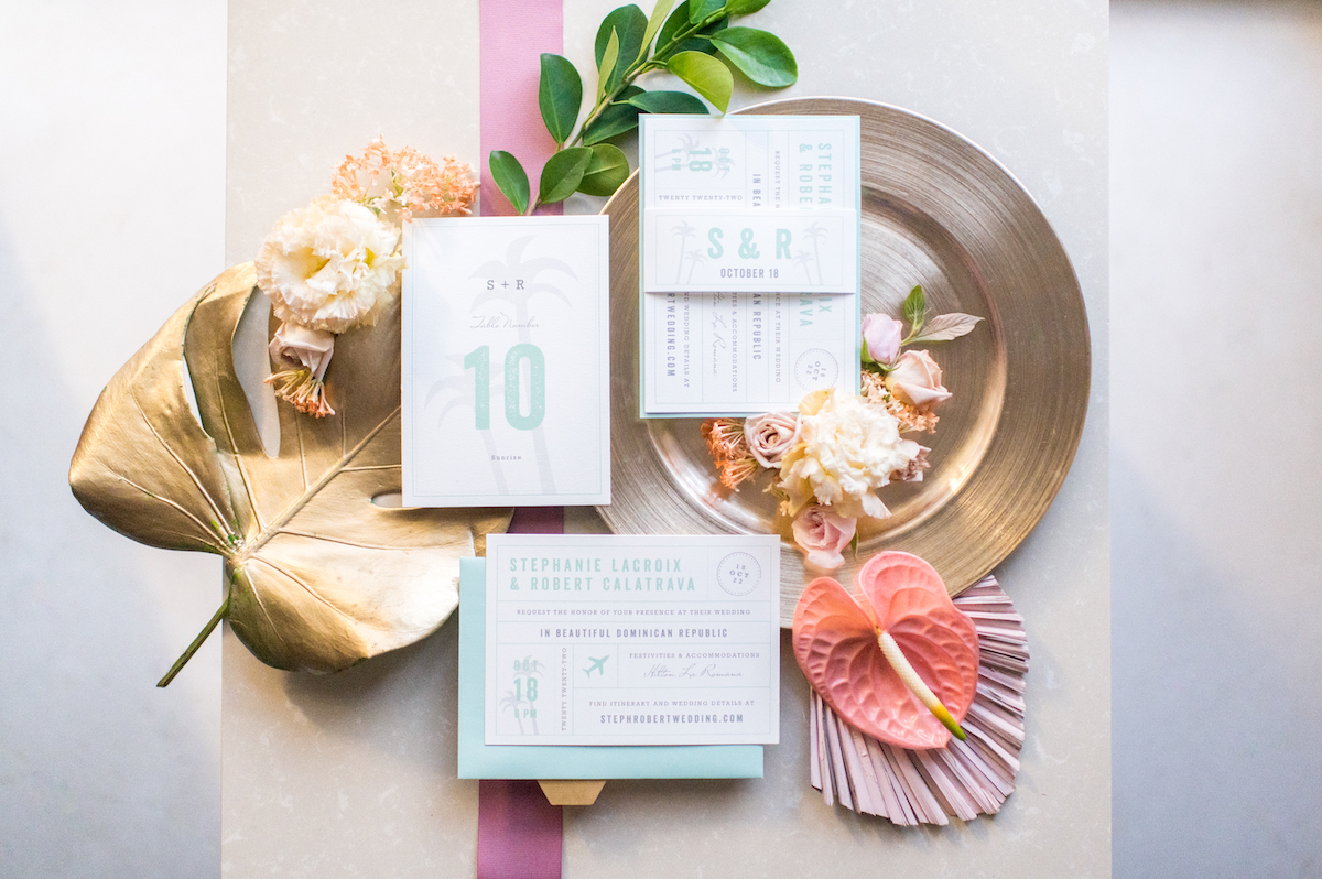 Elegant tropical-themed wedding invitations displayed with pink and white flowers, a gold leaf, and a decorative plate on a light surface.