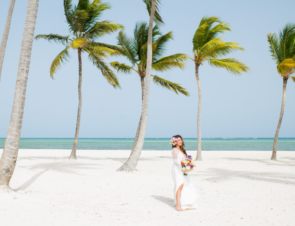 A bride in a white dress stands on a sandy beach with palm trees, holding a bouquet and wearing a flower crown. The ocean is visible in the background under a clear blue sky.