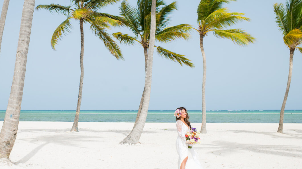 A bride in a white dress stands on a sandy beach with palm trees, holding a bouquet and wearing a flower crown. The ocean is visible in the background under a clear blue sky.