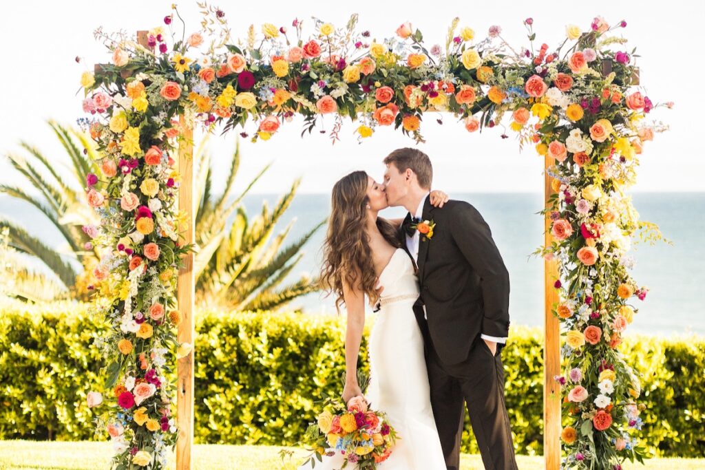 A bride and groom share a kiss under a vibrant floral arch near the ocean, with colorful flowers and greenery surrounding them.