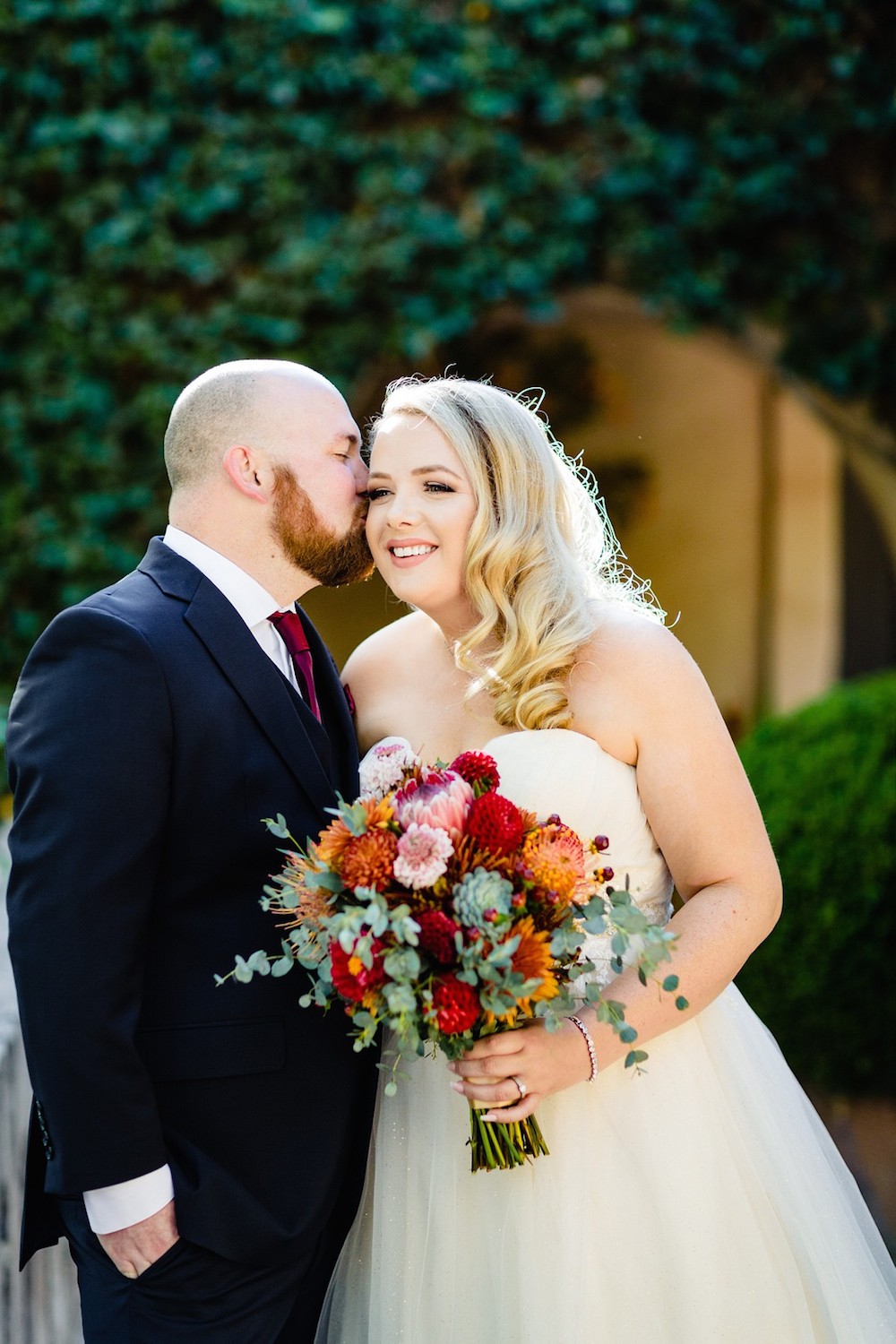 Bride being kissed on the cheek by her groom holding a colorful bouquet of flowers