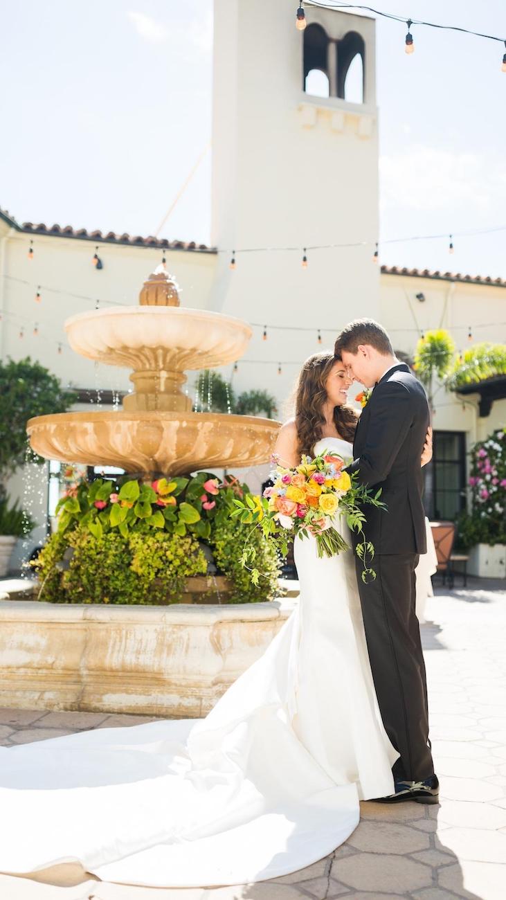 A bride and groom stand close together, holding a bouquet of colorful flowers, in front of a stone fountain at an outdoor venue. The groom wears a black tuxedo, and the bride wears a white dress with a long train.