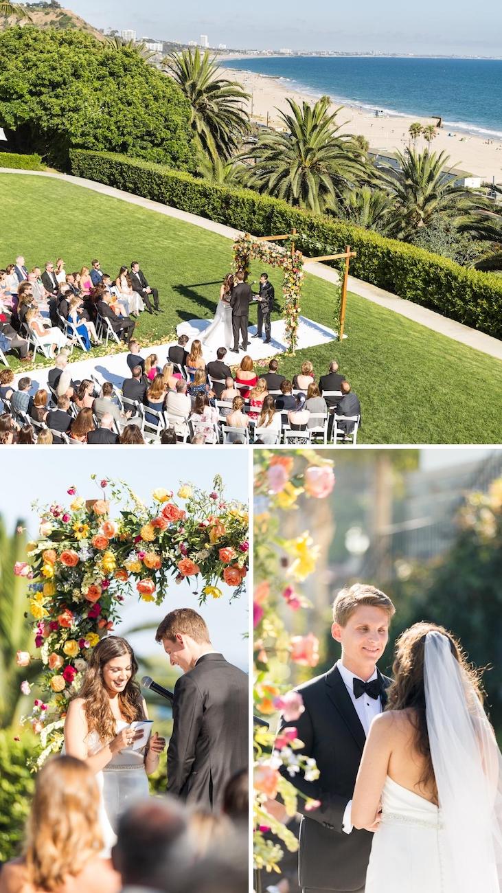 A wedding ceremony taking place outdoors near a beach. The couple stands under a floral arch, with guests seated on white chairs. The background includes palm trees, greenery, and ocean views.