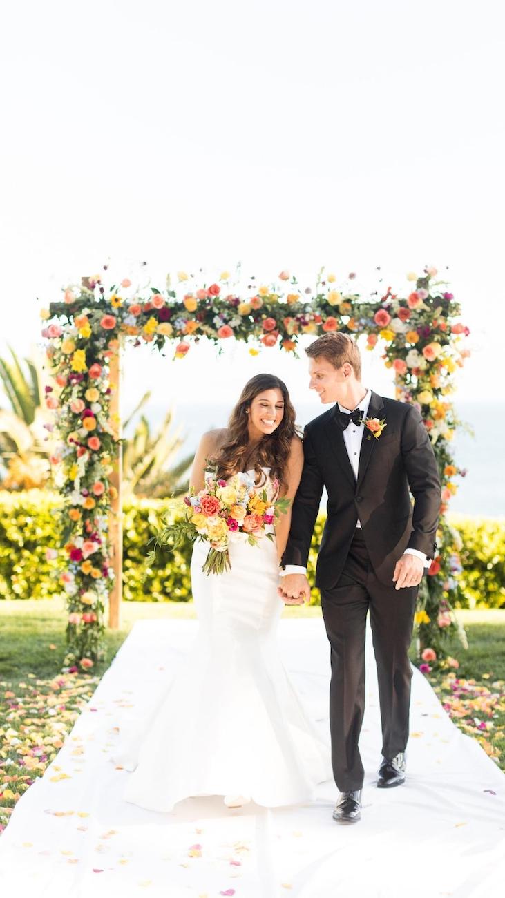 A bride and groom smile as they walk down an aisle outdoors, with a floral arch and greenery in the background. The bride holds a bouquet.