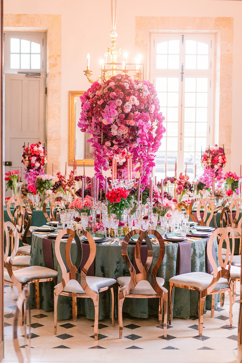 Elegant banquet hall with lavish floral centerpieces, featuring tall arrangements of pink and purple flowers, surrounded by round tables set with china, glassware, and rose gold chairs.