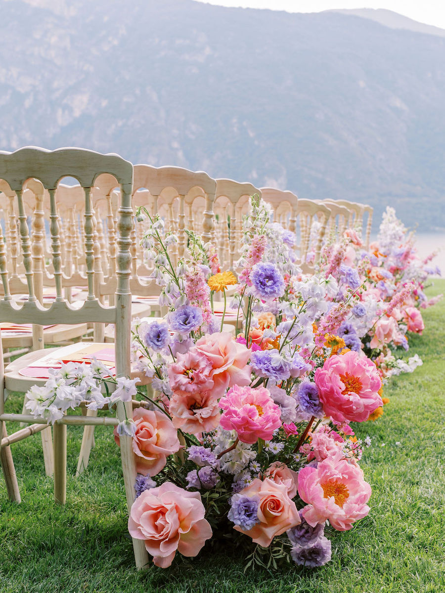 Rows of wooden chairs with colorful floral arrangements of pink, purple, and white flowers along the aisle on a grassy area with a mountainous backdrop.