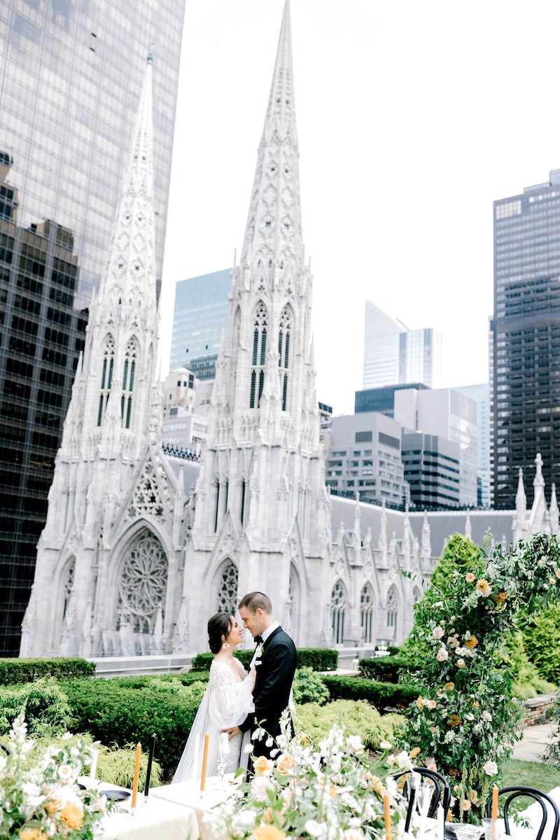 couple surrounded by orange and white florals with a white church with two steeples in the background