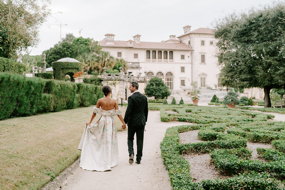 bride and groom walk away from camera in a garden leading to an old stone building