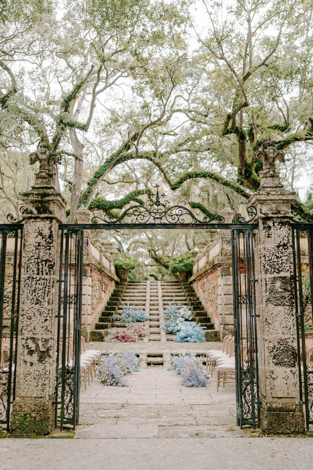 A wrought iron gate leads to a stone staircase overflowing with colorful flowers for a picturesque outdoor wedding ceremony.