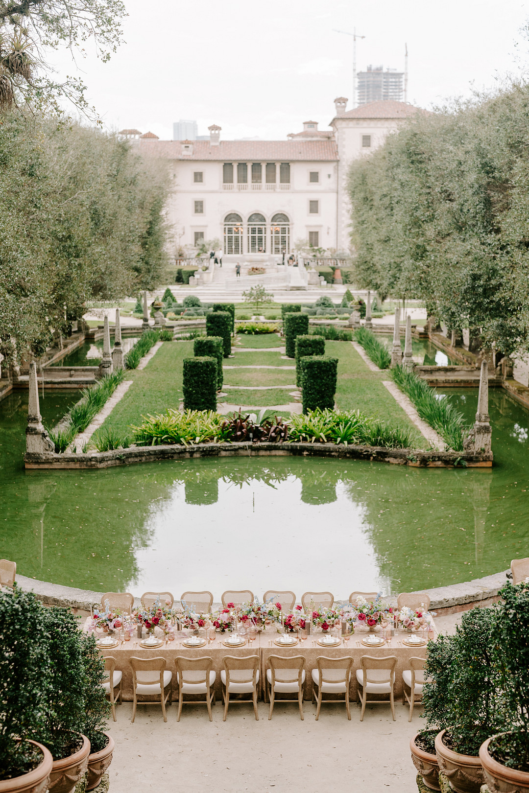 A long wedding reception table with white tablecloths and floral centerpieces set in front of a grand mansion with a pond in the background.