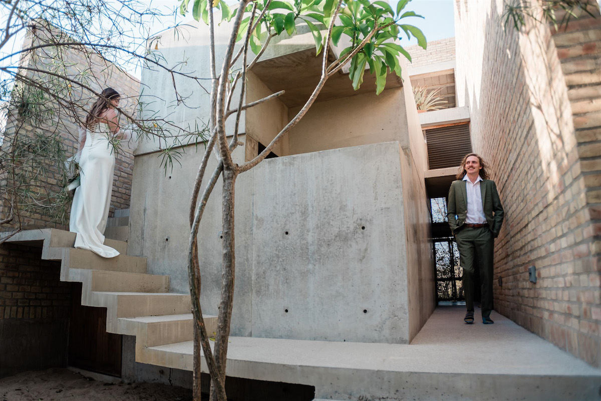 A woman in a white dress stands on outdoor concrete stairs while a man in a suit stands below near a modern concrete and brick building with trees around.
