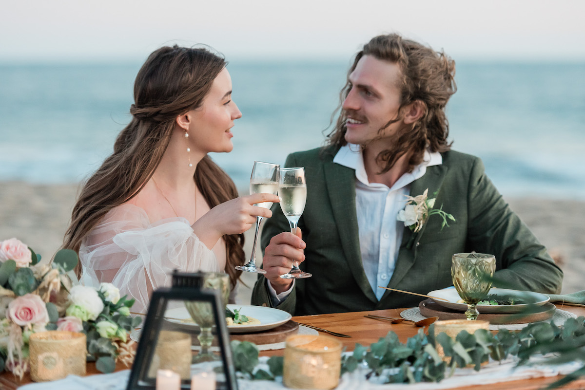 A couple dressed formally sits at a decorated table on the beach, smiling and toasting with glasses of white wine.