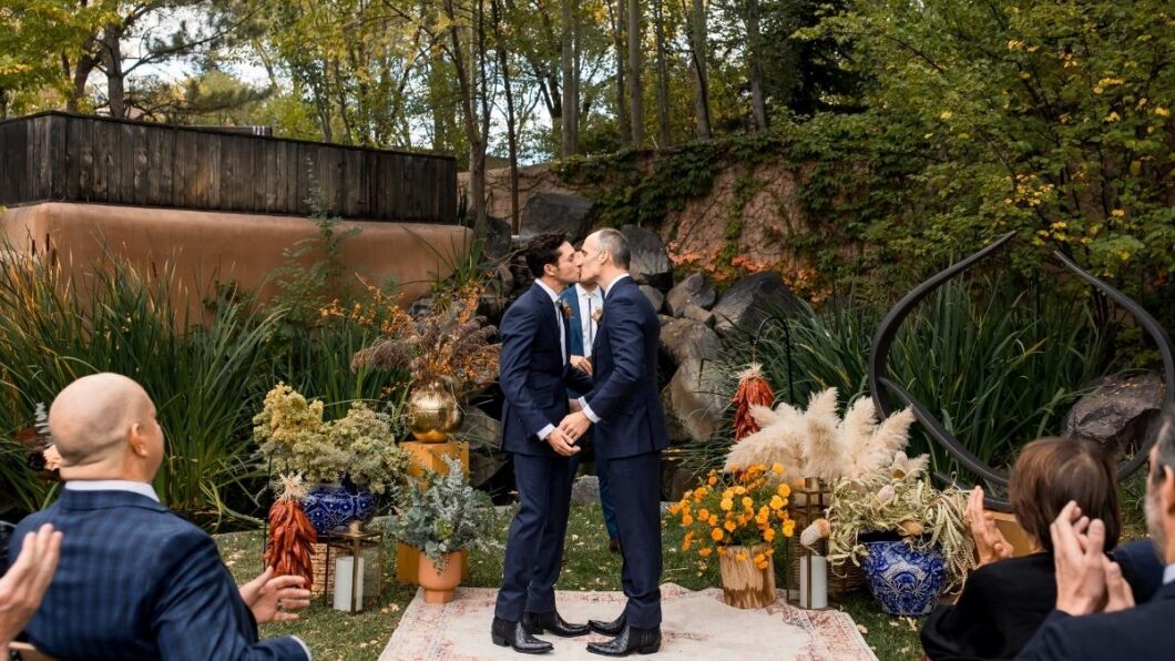 Two men in suits kiss at an outdoor wedding ceremony, standing on a rug surrounded by plants and decorative vases, with greenery and trees in the background.
