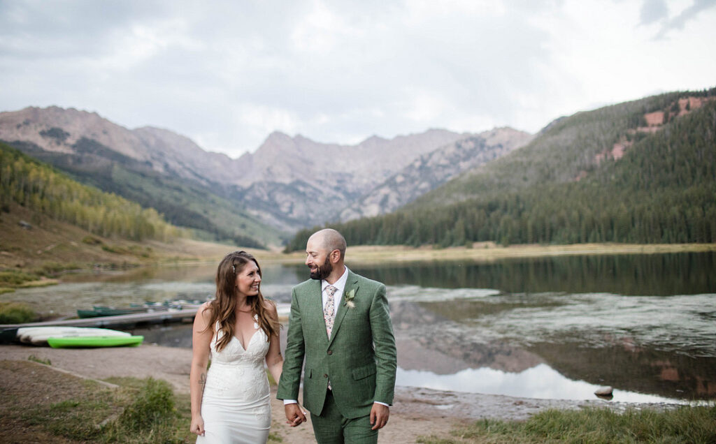 a bride and groom smile at each other as they walk away from a lake surrounded by a jagged mountain range