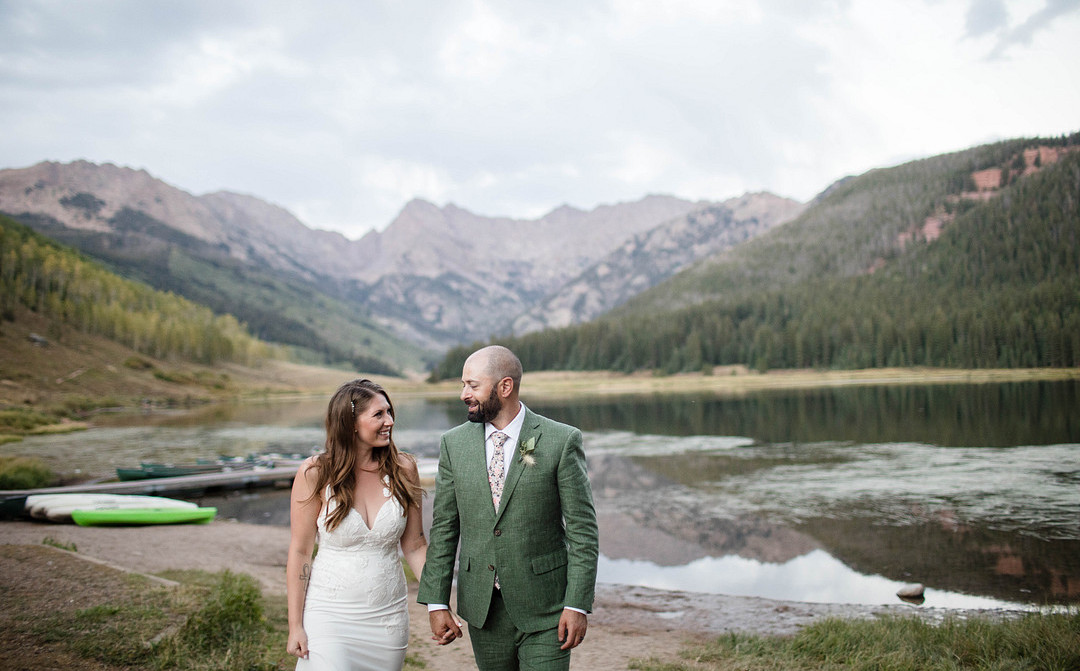 a bride and groom smile at each other as they walk away from a lake surrounded by a jagged mountain range