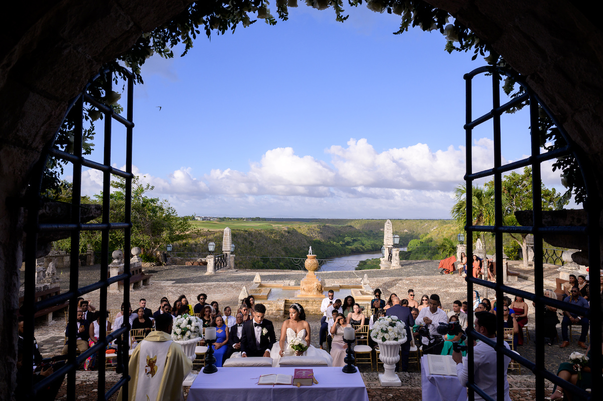 bride and groom kneel with priest; guests sit in gold chairs behind with a backdrop of a Dominican river