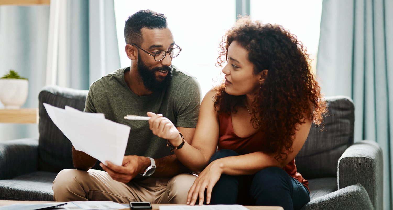 A man and woman sitting on a couch, holding papers and a pen, appear to be discussing documents together in a living room.