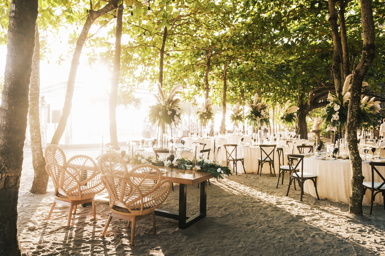 Wedding reception of tables and rattan chairs on a beach under trees