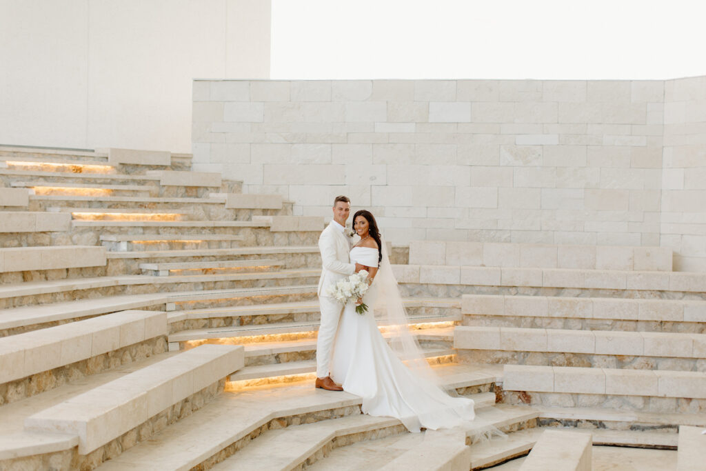 A bride and groom pose together on illuminated stone steps in front of a white stone wall. The bride holds a bouquet and wears an off-the-shoulder dress with a train and veil.