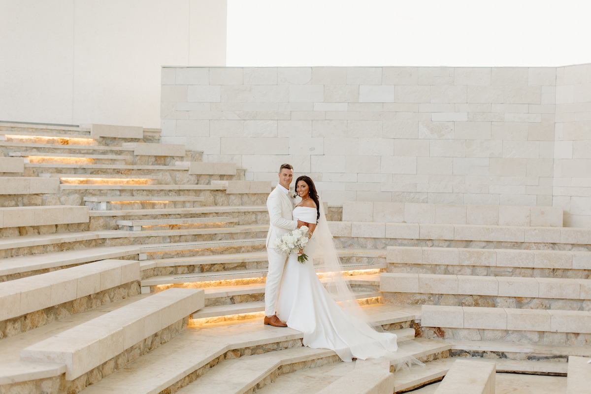 A bride and groom pose together on illuminated stone steps in front of a white stone wall. The bride holds a bouquet and wears an off-the-shoulder dress with a train and veil.
