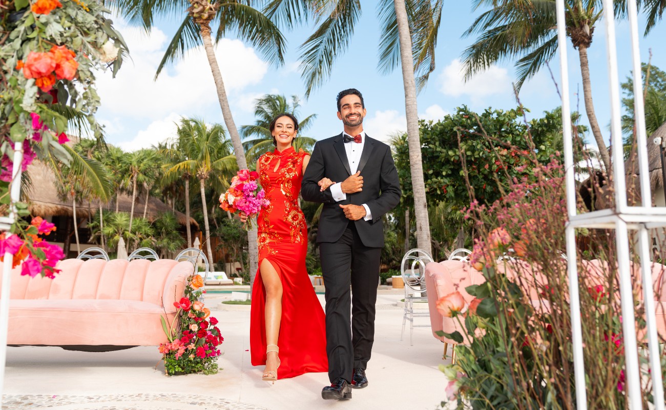Bride wearing a red dress walking with her groom along a wedding reception set up on beach with pink florals
