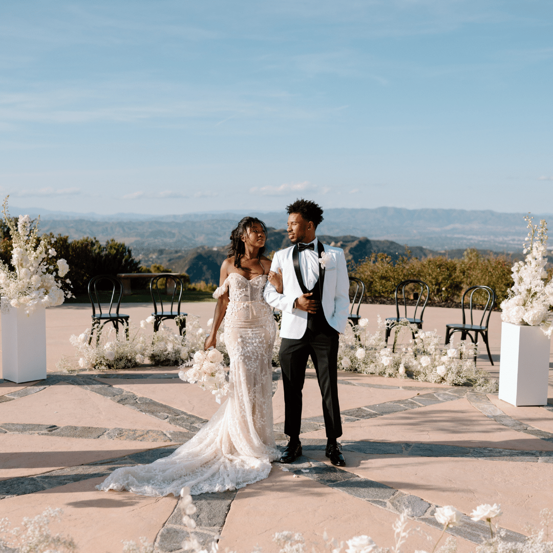 A couple dressed in wedding attire, the bride in a gown and the groom in a white jacket and black pants, walk together on an outdoor patio with chairs and floral arrangements in the background.