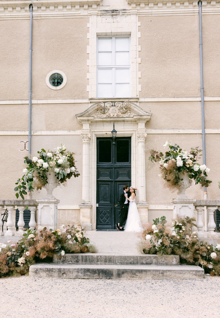 Married couple on steps of French Chateau surrounded with flowers. 